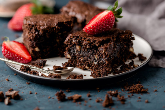 two pieces of chocolate brownie,closeup, on saucer with slices of strawberries, crumbs. Dark blue background