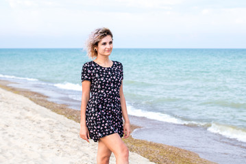 beautiful female with curly hairs in black dress in flowers is standing near water on the beach. Orange sunset light on the face