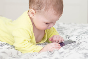 Little child with a cell phone. Baby 7 months interested in the smartphone, lies on the bed and pokes a finger at the screen of the smartphone.