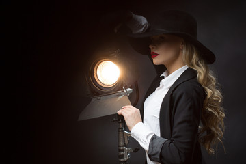 Side view of attractive model in black hat, suit and white shirt standing near lamp and looking aside in studio. Sexy blonde with red lips and long curls posing. Concept of sensuality and beauty.