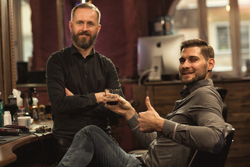 Satisfied male client of barbershop sitting in chair, drinking whiskey, looking at camera and showing sign ok. Handsome man with new haircut liking trendy hairstyle. Concept of professional barber.