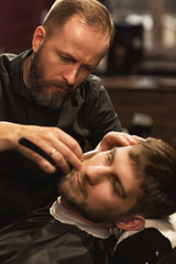 Fototapeta premium Selective focus of careful barber wearing black shirt in process of working in barbershop. Serious male master shaving beard of young client with razor. Concept of beard shape and care.