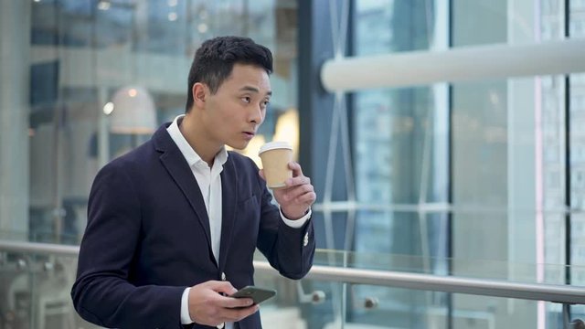 Middle Shot Slow Motion Of Asian Young Businessman Black Hair Dressed In Suit Drinking Coffee Holding Phone Walking In Business Center 