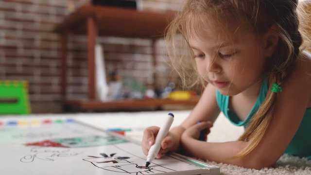 Video Shot of happy little girl lying and drawing with felt-tip pen on white capet at home. Childhood concept. Studio video shot of child's daily life in 4K definition.