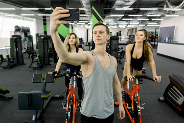 Fototapeta na wymiar Handsome young man couch making selfie on the phone in gym, while two young pretty girls training on exercise bikes together in fitness club