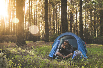 Beautiful young male drinking a cup of coffee in camping tent. Tourist boy enjoying outdoor life in mountain On background trees and setting sun Tourism adventure active lifestyle and healthy concept