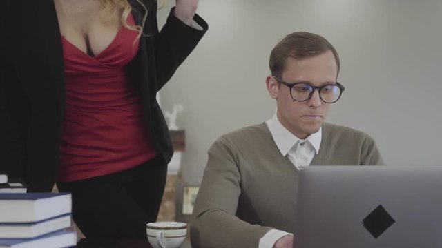 Portrait young modestly dressed man in glasses sitting at table at home, working on laptop. Beautiful woman with a deep neckline bends over and offers him coffee, a man is confused. Cute nerd and hot