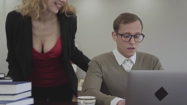 Portrait young adorable modestly dressed man in glasses sitting at table at home, working on laptop. Beautiful woman with deep neckline bends over and looking on screen, a man is confused. The nerd