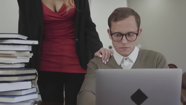 Young modestly dressed man in glasses sitting at table at home, working on laptop. Beautiful woman with deep neckline bends over and putting hand on his shoulder. The nerd trying to shake off the hand