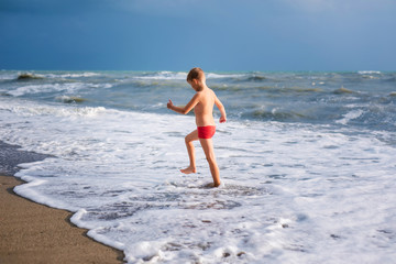 Blonde boy running and jumping on the beach on blue sea shore in summer vacation at the day time. Blue ocean with white big wawes on the background