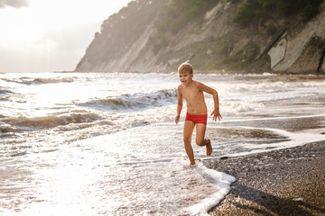 Blonde boy running and jumping on the beach on blue sea shore in summer vacation at the day time. Blue ocean with white big wawes on the background