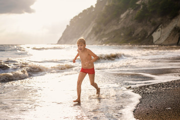 Blonde boy running and jumping on the beach on blue sea shore in summer vacation at the day time. Blue ocean with white big wawes on the background