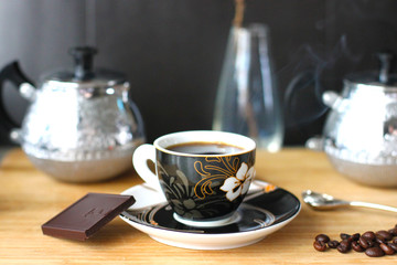 A Cup of steaming black coffee on a saucer with a spoon and coffee beans on a black background with Melchior sugar bowl and milk jug.