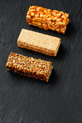 Cereal granola bar with peanuts, sesame and sunflower seeds on a cutting board on a dark stone table. View from above. Three Assorted Bars