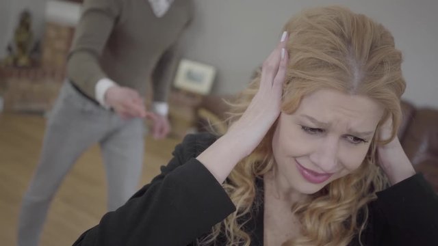 Pretty blond woman in the foreground covering her ears, an emotional angry man yelling and screaming on the background. The couple has a quarrel. Difficulties in relationship