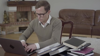 Portrait young modestly dressed man in glasses sitting at the wooden table in the office working on his laptop. Unrecognizable angry woman bringing and throwing on the desk stack of books