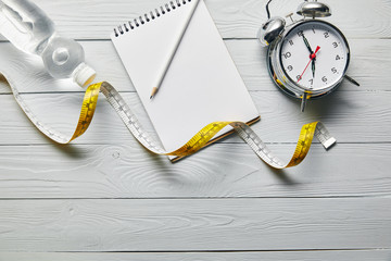 top view of measuring tape, blank notebook, water and alarm clock on wooden white background with...