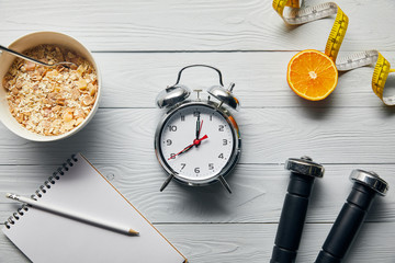 top view of silver alarm clock, notebook with pencil, breakfast cereal, measuring tape, dumbbells...