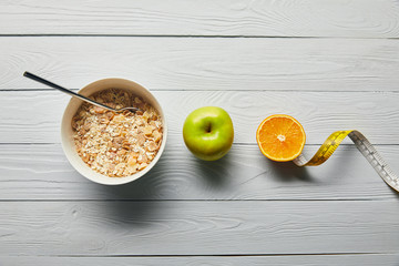 flat lay with breakfast cereal in bowl, apple, orange and measuring tape on wooden white background