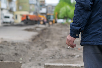 Hand man with a cigarette on the background of the destroyed and repair the road in the yard of a house with construction machinery and machines