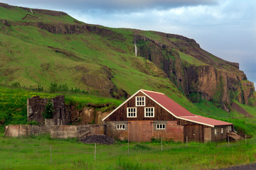 Fototapeta na wymiar The wooden house with red roof back of the fences under the green rocky cliffs surrounded with green grasses