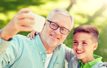 family, generation, technology and people concept - happy grandfather and grandson with smartphone taking selfie at summer park