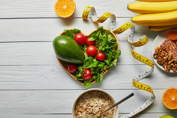 top view of fresh fruits, vegetables in heart-shaped bowl, measuring tape and cereal on wooden white background