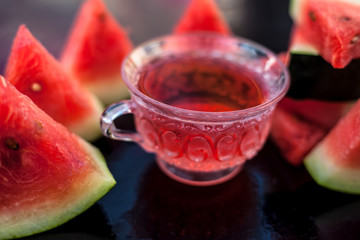 Fruit ice tea of watermelon seeds in a transparent glass cup on wooden surface along with triangular pieces of watermelon.