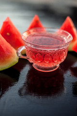 Vertical shot of fruit ice tea of watermelon seeds in a transparent glass cup on wooden surface along with triangular pieces of watermelon.
