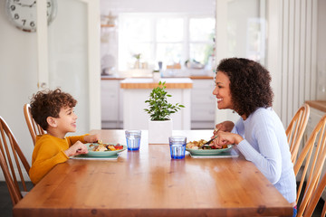 Single Mother Sitting At Table Eating Meal With Son In Kitchen At Home