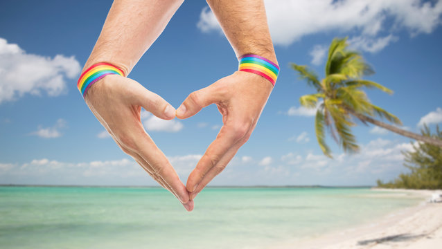 lgbt, same-sex love and homosexual relationships concept - close up of male couple hands with gay pride awareness wristbands showing heart gesture over tropical beach background in french polynesia