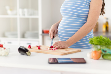 Obraz na płótnie Canvas pregnancy, cooking food and healthy eating concept - close up of pregnant woman with kitchen knife chopping radish on wooden cutting board at home