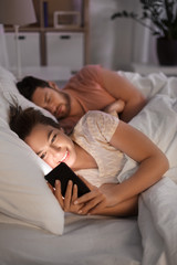 Obraz na płótnie Canvas technology, internet addiction and cheat concept - happy smiling woman using smartphone at night while her boyfriend is sleeping