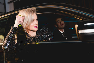 stylish man and beautiful young woman in formal wear with champagne bottle sitting in car