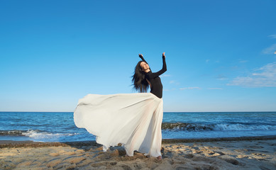 Classical dancer woman. Charming ballerina with windy hair in white chiffon skirt dancing by the sea background. Sun shines on her. Horizontal.