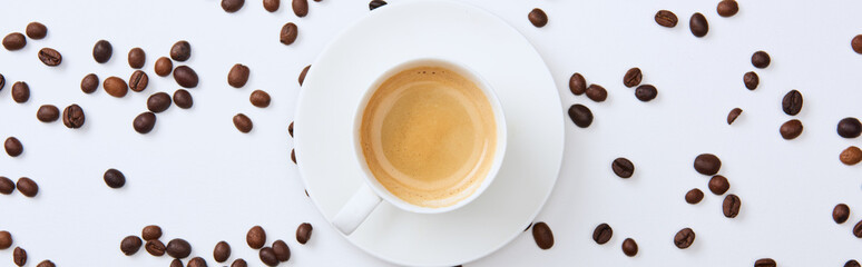 panoramic shot of delicious coffee in cup near scattered roasted beans on white background