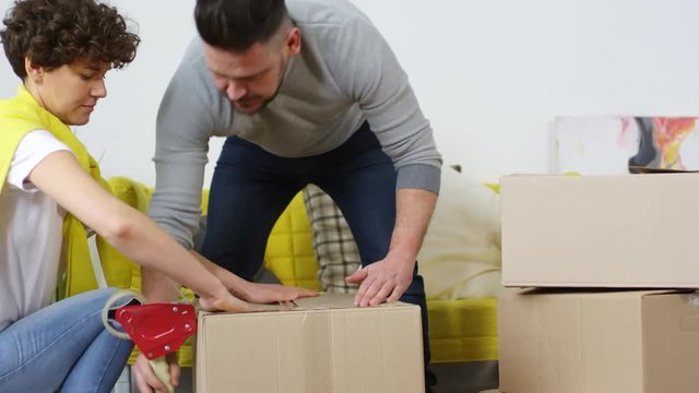 Medium shot of middle-aged Caucasian husband and wife settling down together in their new home after relocation and taping over cardboard boxes to put away for storage