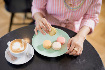 Fototapeta na wymiar Top view of mixed race woman in pink striped dress holding cookie while sitting in pastry shop. On table are coffee and plate with cookies.