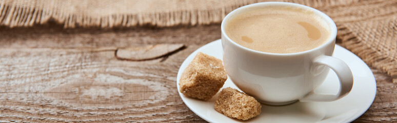 panoramic shot of delicious coffee with foam in cup on saucer with brown sugar near sackcloth on wooden background