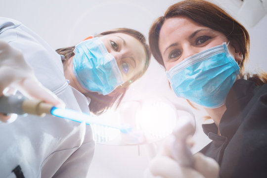 Bottom view of two women dentists in surgical mask holding tools and looking at camera. Patient point of view to dentist.