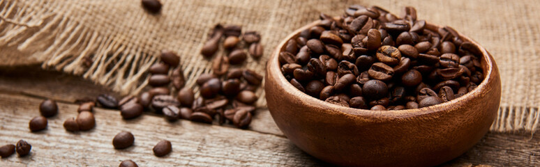 close up view of roasted coffee beans in bowl near sackcloth on wooden board, panoramic shot