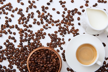 top view of coffee in cup on saucer near scattered roasted beans, wooden bowl and milk jug