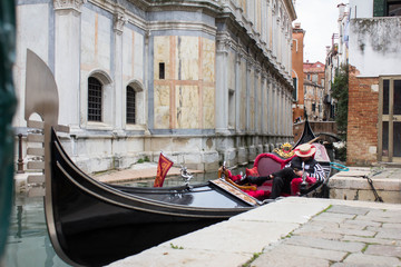 Huge long gondola moored to the narrow canal embankment and gondolier in traditional Venetian striped clothes and wearing the hat sitting in a boat. Typical Italian architecture of residential facades