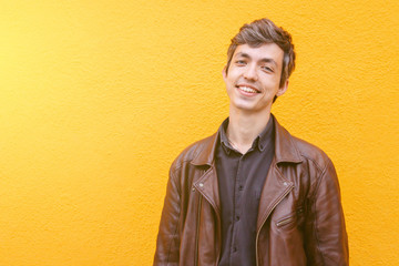 Happy handsome greyish young guy staying against the yellow wall of the residential building. Smiling european man in a jacket. Casual happy student in leather jacket. Portrait of the young man.