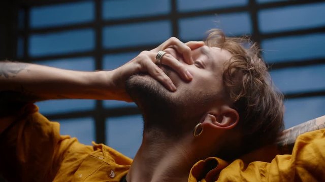 Close up of stylish Caucasian man modern movement performance dancing in darkness time, tattoo on hands with rings touching head and face of guy freestyle moving dressed in orange shirt and earrings