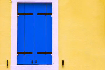 Bright yellow painted wall of residential building and wooden old shutter window with beautiful traditional blue shutters. Copy space and space for inscription on the right. Cute residential houses.