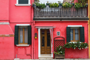 Fototapeta na wymiar Red painted wall of residential building with wooden entrance door and shutter windows with old traditional shutters. Cute flowers and plants on window sills and flowers on the balcony.