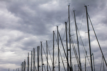 A row of long yacht masts against the backdrop of rainy gray dramatic sky. Mooring of boats, navigation, storm, dangerous weather conditions, strong wind on the waterfront. Thick clouds on the sky.
