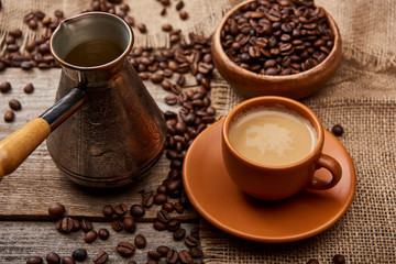 coffee beans in bowl and cezve near cup of coffee on wooden background