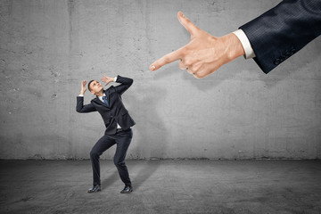 Little businessman covering head with hands and looking up at big man's hand pointing finger-gun at...